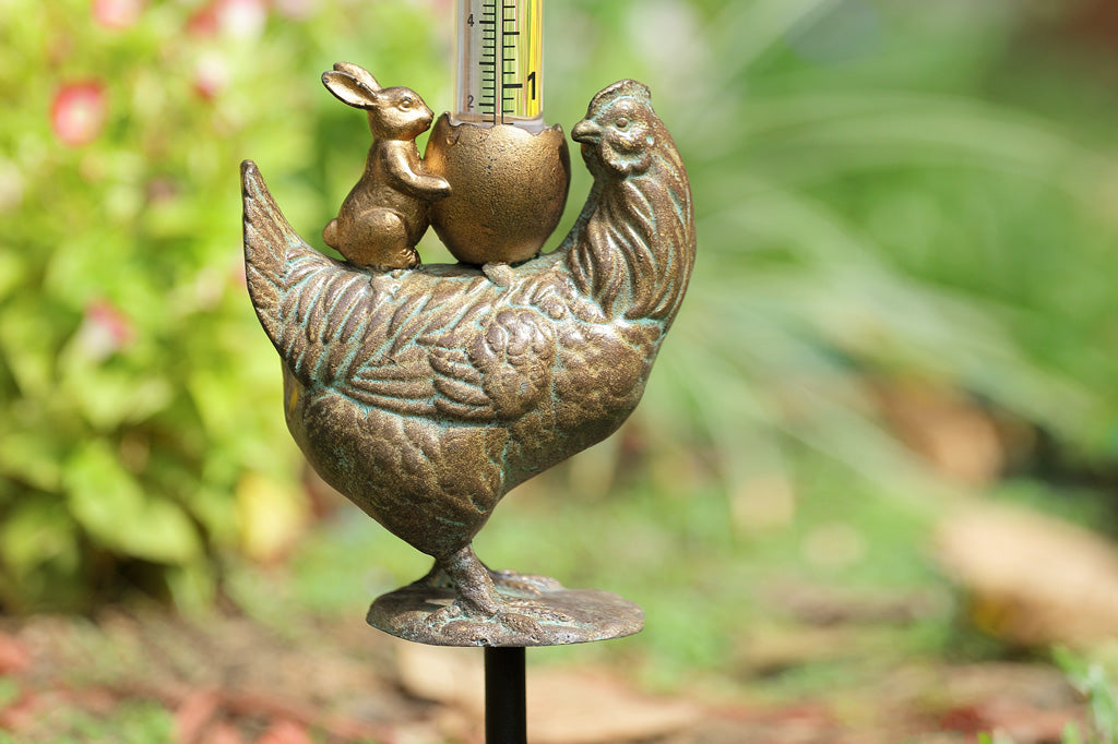 Closeup view of cast metal chicken with bunny and cracked egg with rain gauge, set in garden bed  