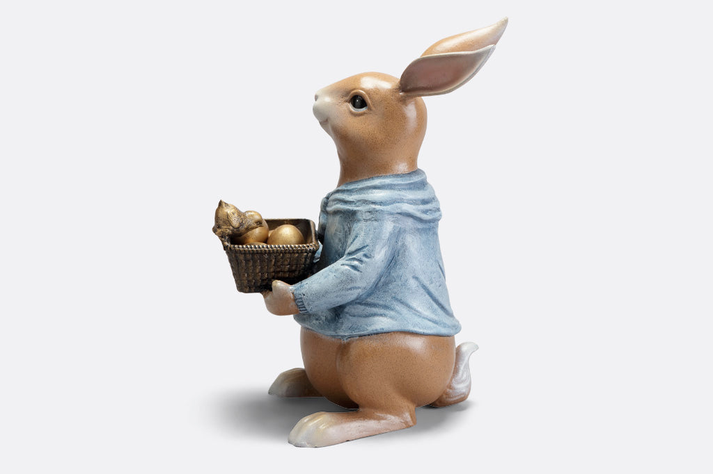 side view of cast metal sculpture of storybook-style rabbit in a blue shirt, holding a basket of four removable golden eggs. A chick perches on edge of basket to get a closer look.