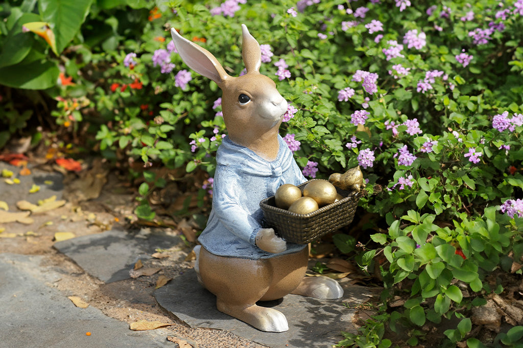 Shown on floral garden path,  cast metal sculpture of storybook-style rabbit in a blue shirt, holding a basket of four removable golden eggs. A chick perches on edge of basket to get a closer look.