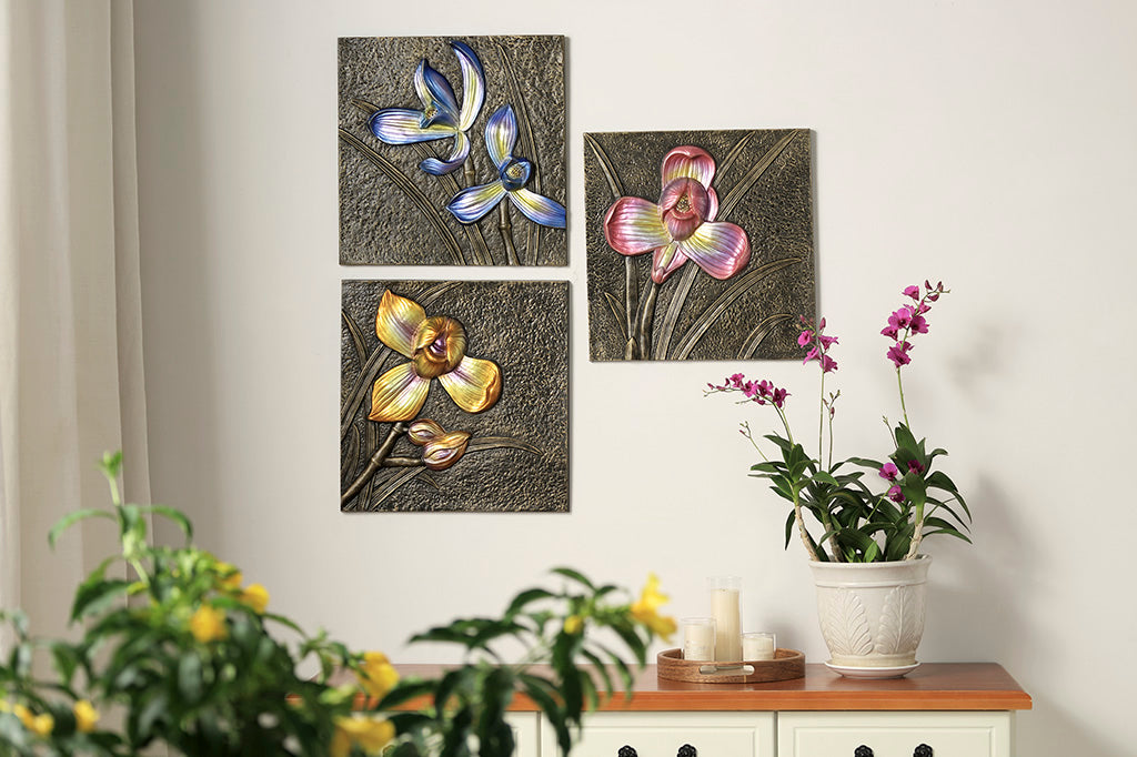 Three bronze decorative wall tiles with metallic orchid motifs in blue, violet-pink, and yellow, shown hung above a desk with candles and potted orchid