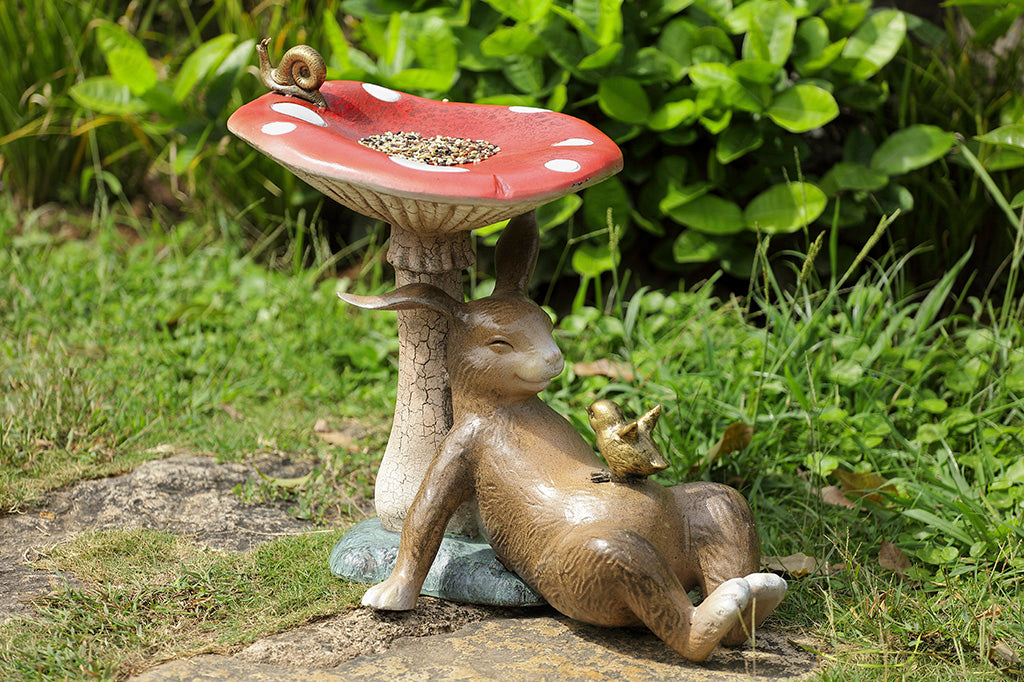 Bunny under a toadstool sculpture with chick and snail - mushroom is birdbath/birdfeeder, shown in garden