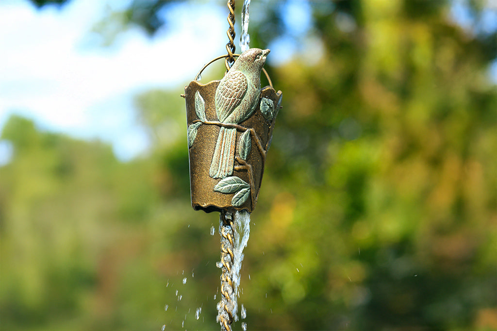 water cascades through a cast metal rain cup attached to an outdoor rain chain. Rain cup features a finch in verdigris patina on a branch and leaf design.