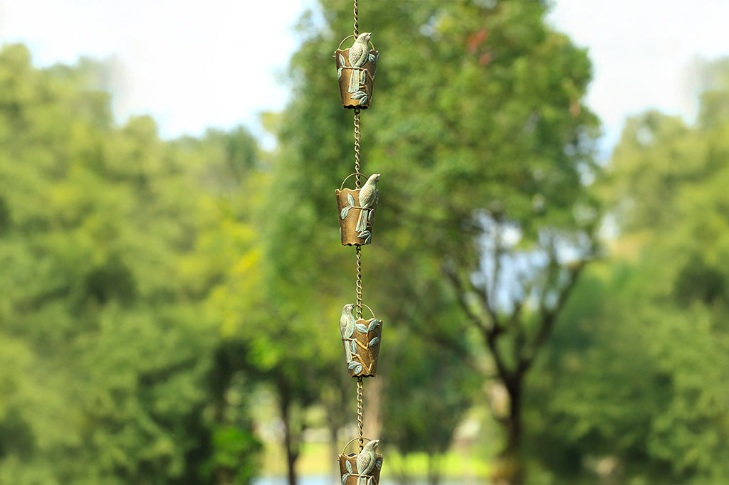 8 rain cups attached to an outdoor rain chain shown outside. Rain cup features a finch in verdigris patina on a branch and leaf design.