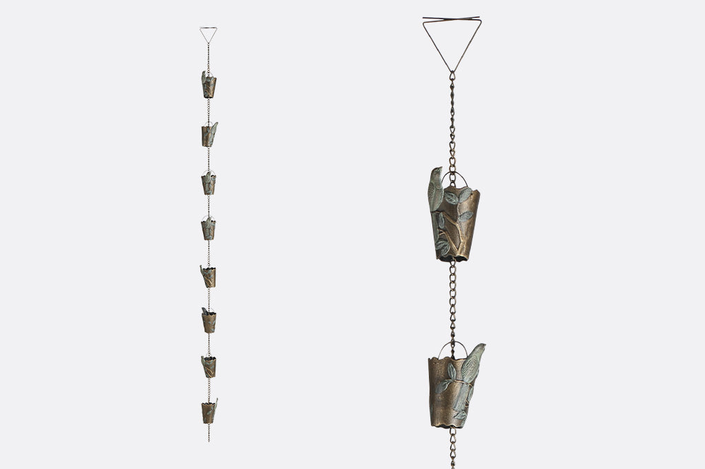 8 rain cups attached to an outdoor rain chain shown outside. Rain cup features a finch in verdigris patina on a branch and leaf design.