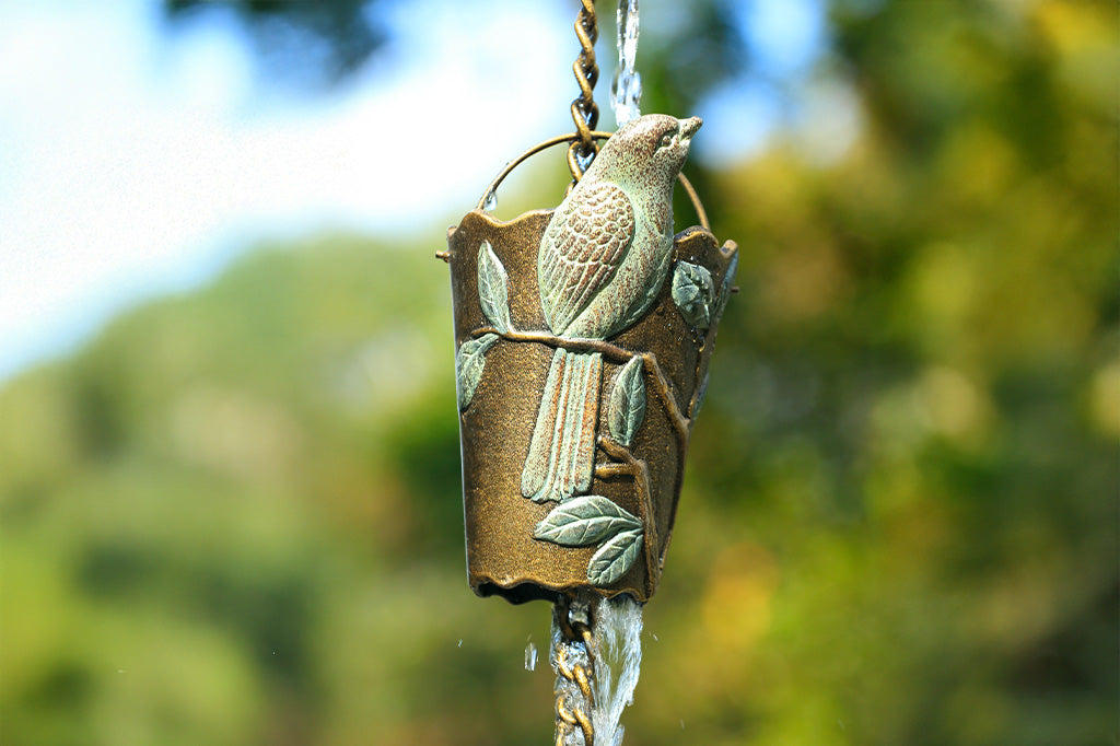 Closeup detail of rain chain's rain cup featuring a finch in verdigris patina on a branch and leaf design.