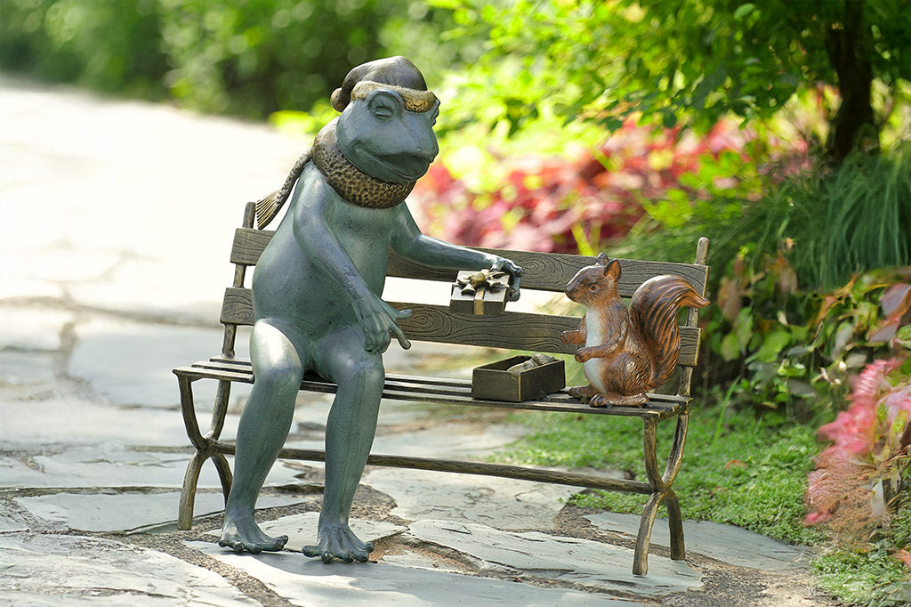 Decorative statue of a frog sharing a box of acorns with a with a squirrel on a park bench, shown on garden path in an outdoor park