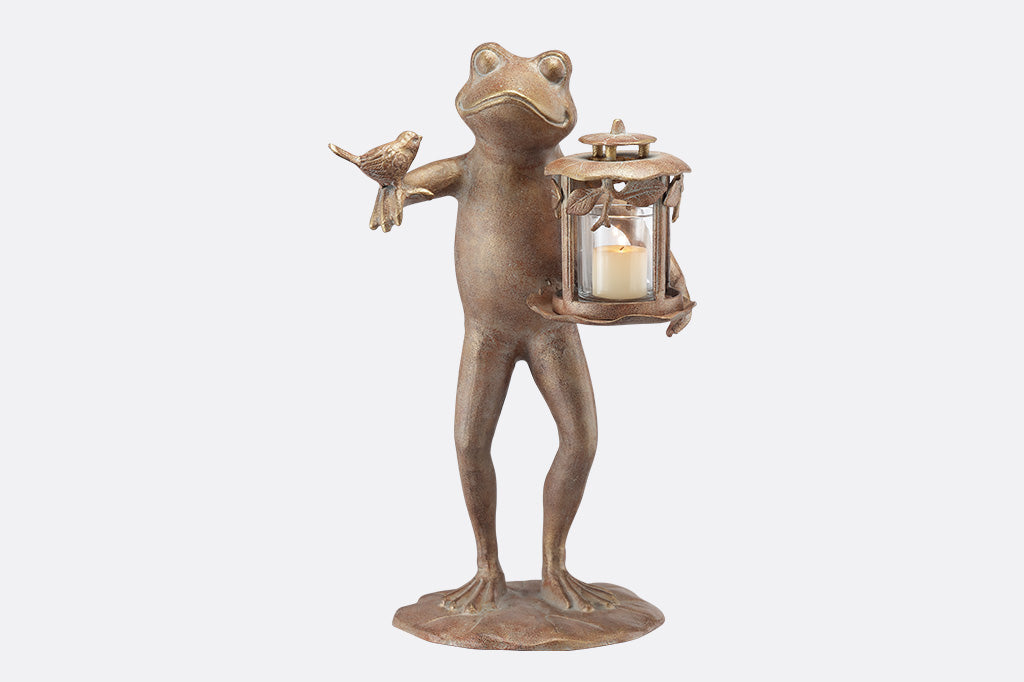 A brown statue of a frog with a bird on its arm and a lantern on its hand against a white background.