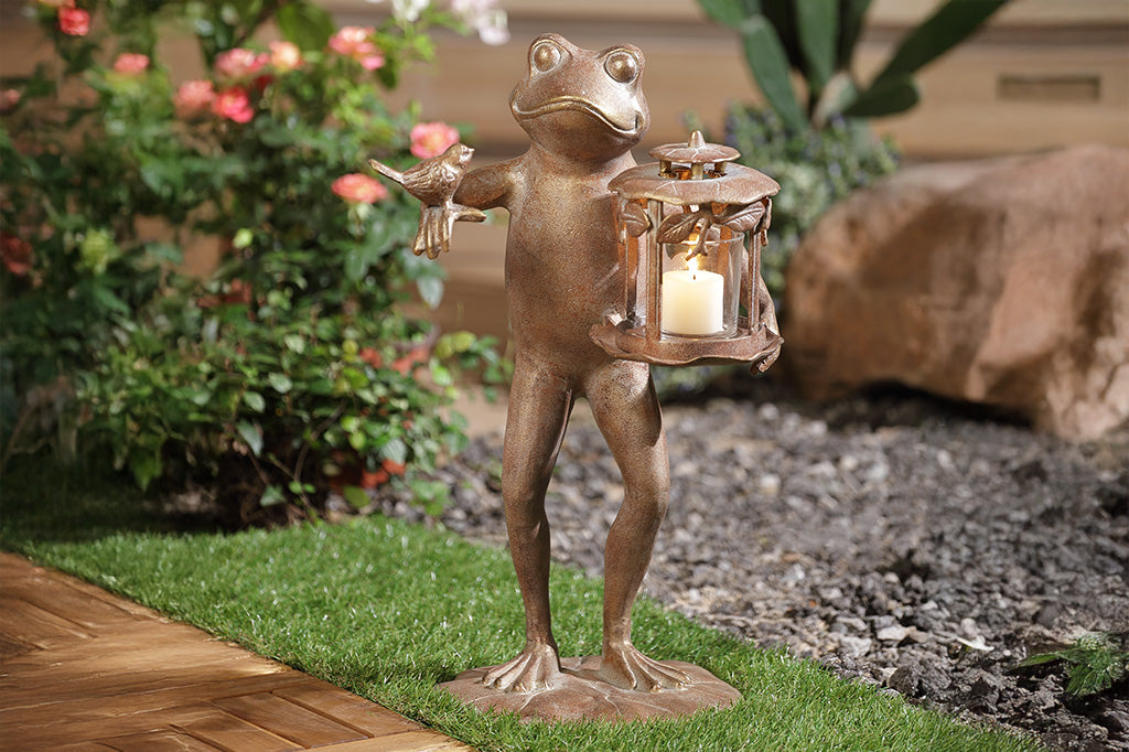 A brown statue of a frog with a bird on its arm and a lantern with a lit candle inside on its hand placed outside in a garden.