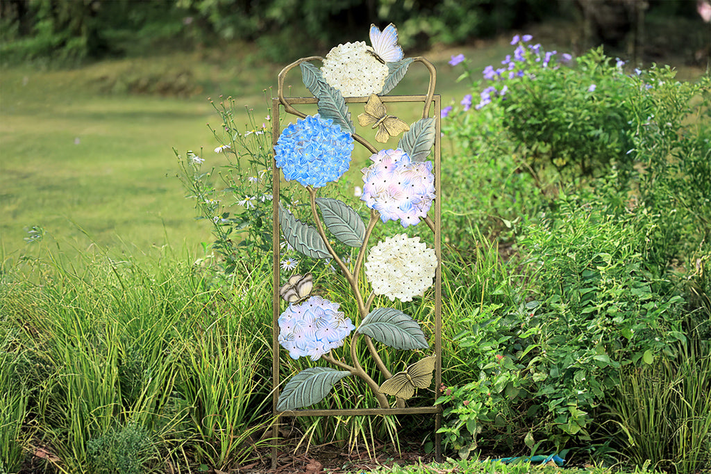 Garden trellis with colored finish on both sides, features white, blue, and purple hydrangea blossoms with multi-hued butterfly accents. Shown in Garden amongst foliage.