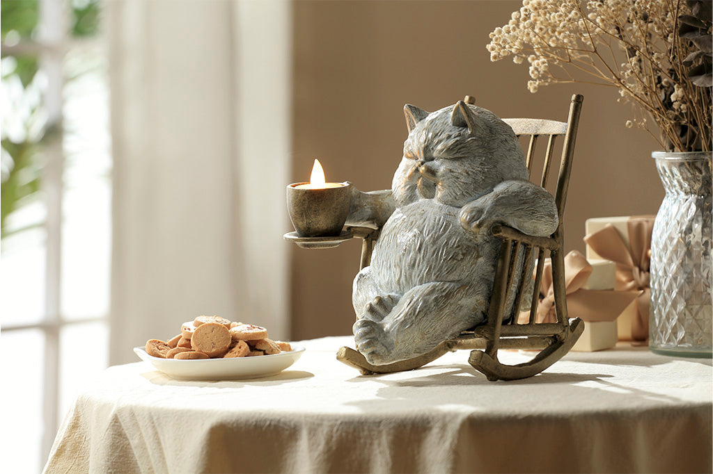 White cat in a rocking chair sculpture. Cat is restfully sitting with eye closes, holding a teacup which holds LED tealight. Placed on table by a window, plate of cookies nearby.