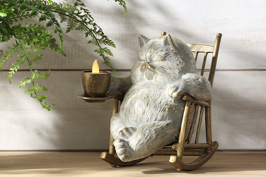 White cat in a rocking chair sculpture. Cat is restfully sitting with eye closes, holding a teacup which holds LED tealight. Placed on wood table near a potted fern