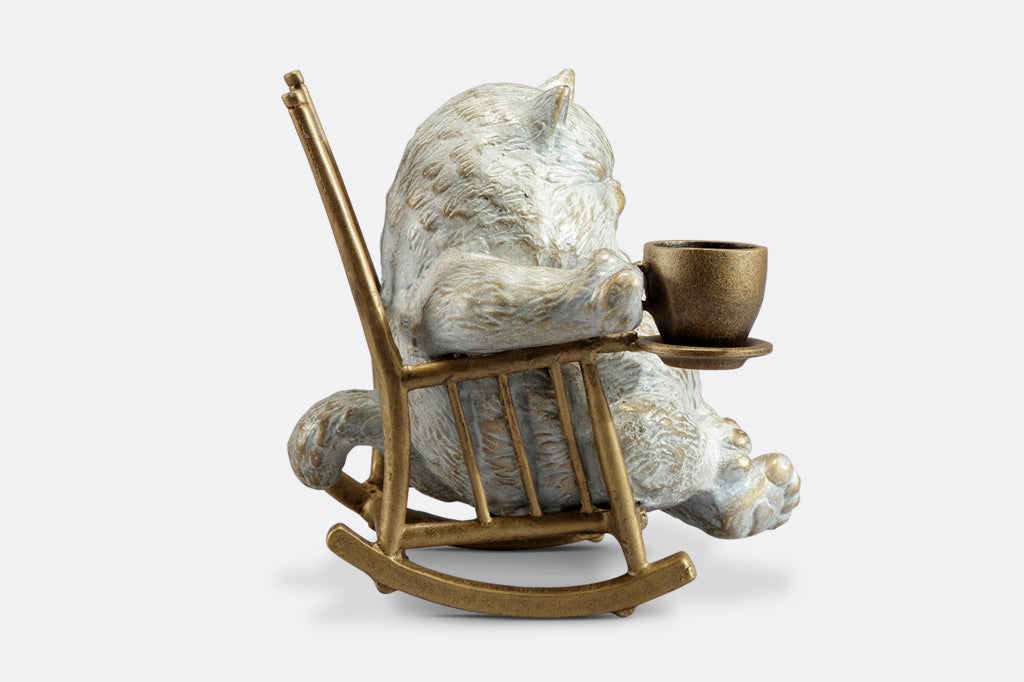 side view of White cat in a rocking chair sculpture. Cat is restfully sitting with eye closes, holding a teacup which could hold LED tealight.