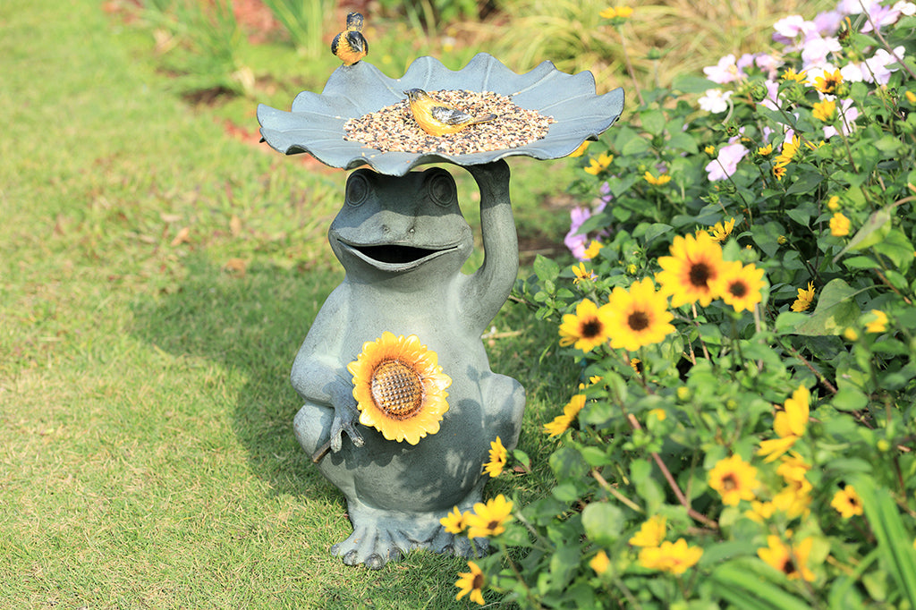 Outdoor birdbath/birdfeeder shown with birdseed in a flower garden. Features a Frog holding a sunflower with yellow songbird accents on a lilypad basin.