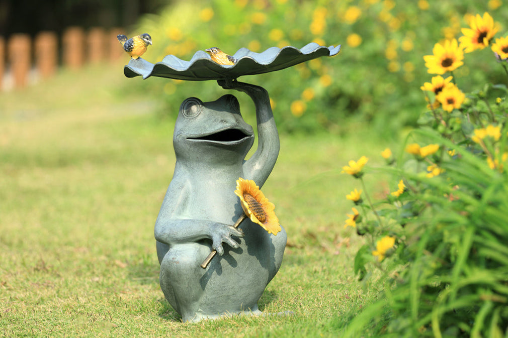 Outdoor birdbath/birdfeeder shown with birdseed in a flower garden. Features a Frog holding a sunflower with yellow songbird accents on a lilypad basin.