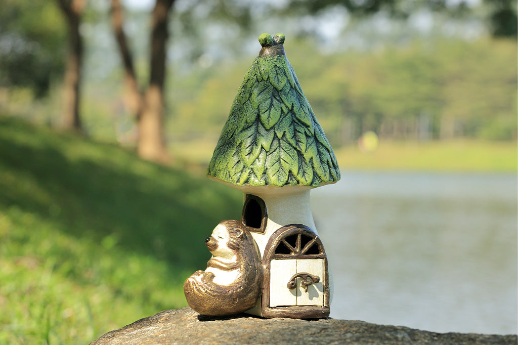 Outdoor Garden candle lantern shows hedgehog sleeping against a woodland house with working door, used to insert votive candle. Shown on rock in garden setting.