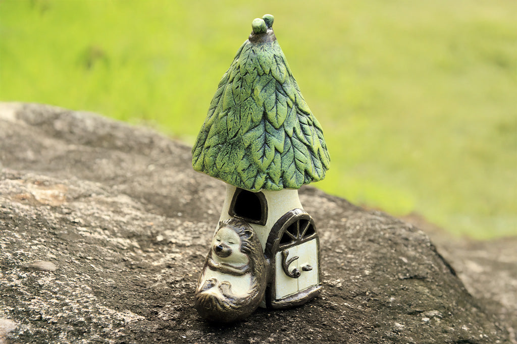 Outdoor Garden candle lantern shows hedgehog sleeping against a woodland house with working door, used to insert votive candle. Shown on rock in garden setting.