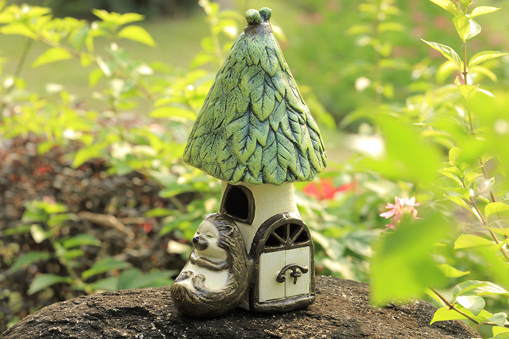 Outdoor Garden candle lantern shows hedgehog sleeping against a woodland house with working door, used to insert votive candle. Shown on rock in garden setting.