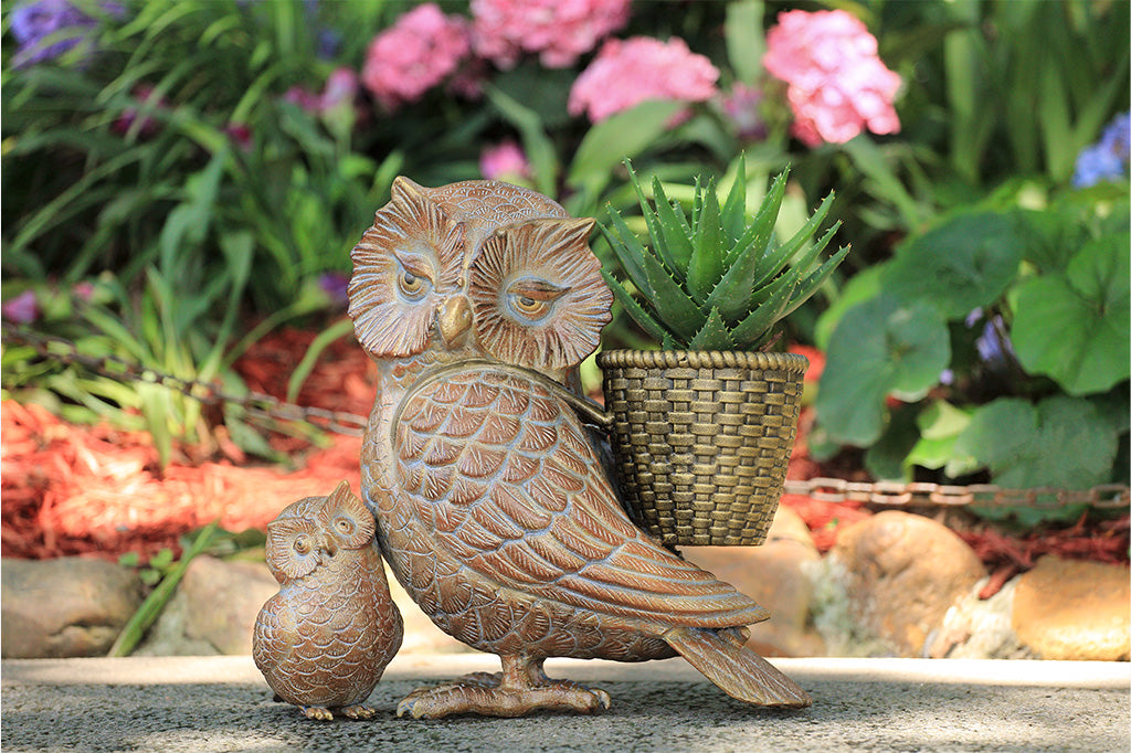 Bronze owl sculpture with a smaller owl figure in front of a potted plant with pink flowers.