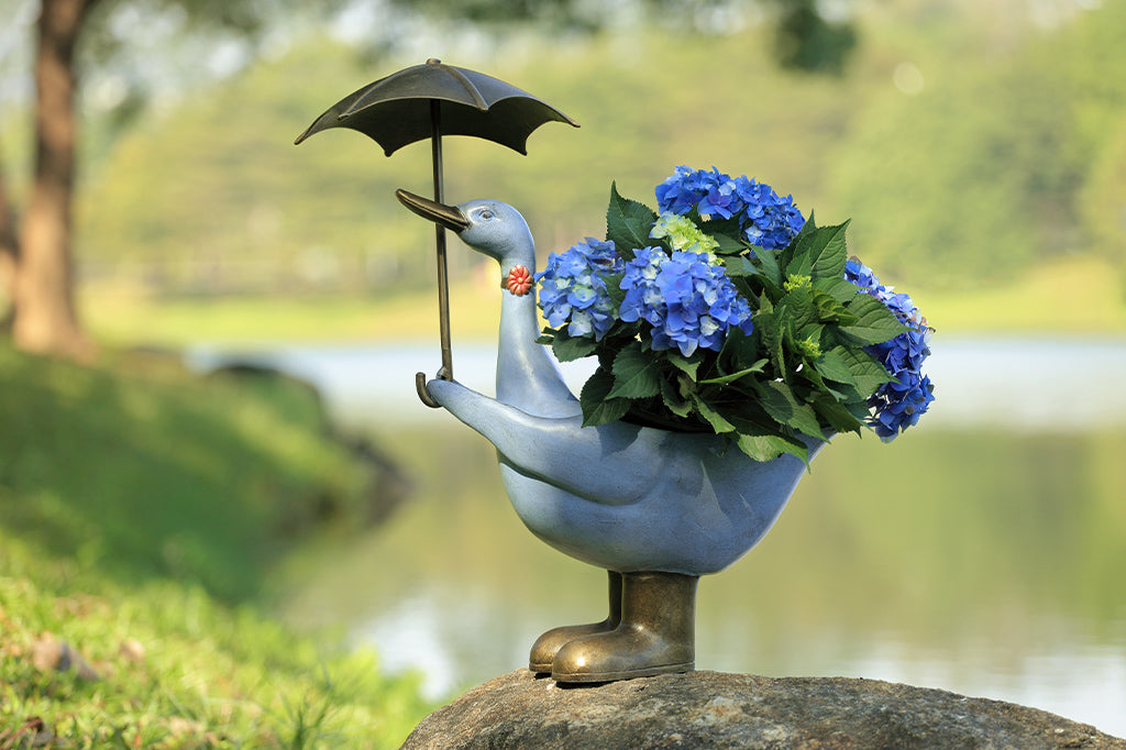 Slate blue goose planter shown holding potted hydrangeas near a lake. She holds an umbrella, flower choker, and rain boots.