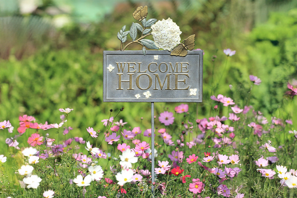 Outdoor Garden Sign with words "Welcome Home" on rectangle frame, adorned with white hydrangea blossom and golden butterflies. Shown in garden with purple flowers.