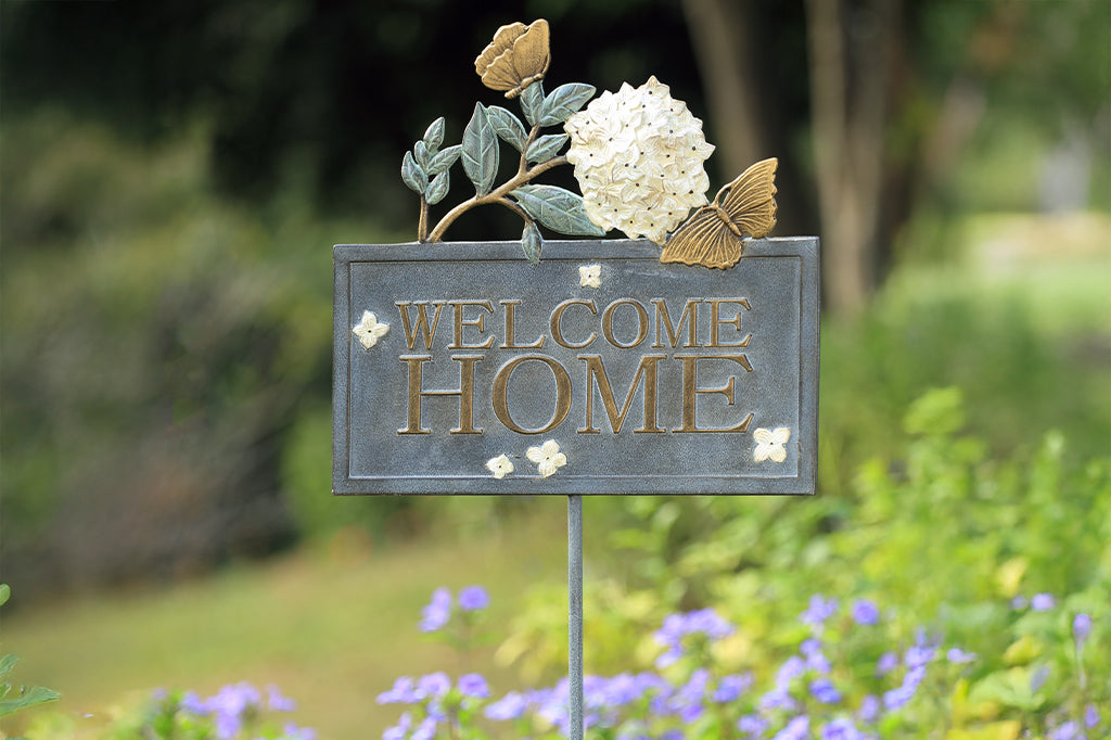 Outdoor Garden Sign with words "Welcome Home" on rectangle frame, adorned with white hydrangea blossom and golden butterflies. Shown in garden with purple flowers.