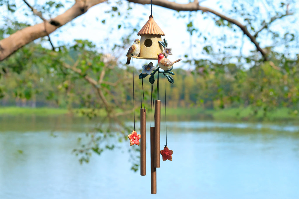 Birdhouse motif cast metal windchime with floral and songbird accents shown hanging from tree by lake. 3 suspended tubes and 2 pendant flower charms make the sounds