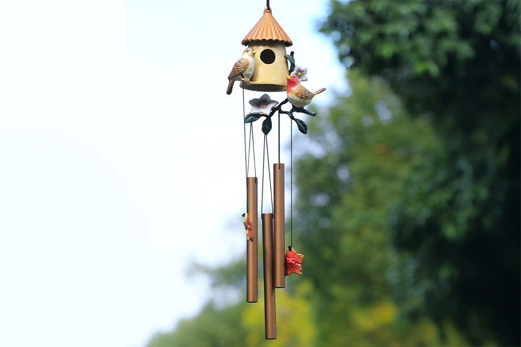 Birdhouse motif cast metal windchime with floral and songbird accents shown hanging from tree by lake. 3 suspended tubes and 2 pendant flower charms make the sounds