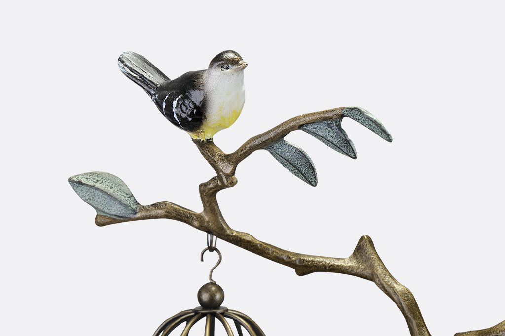 Closeup view of a staked garden accent with branch and leaf motif with songbirds. It supports two hanging candle lantern cages.