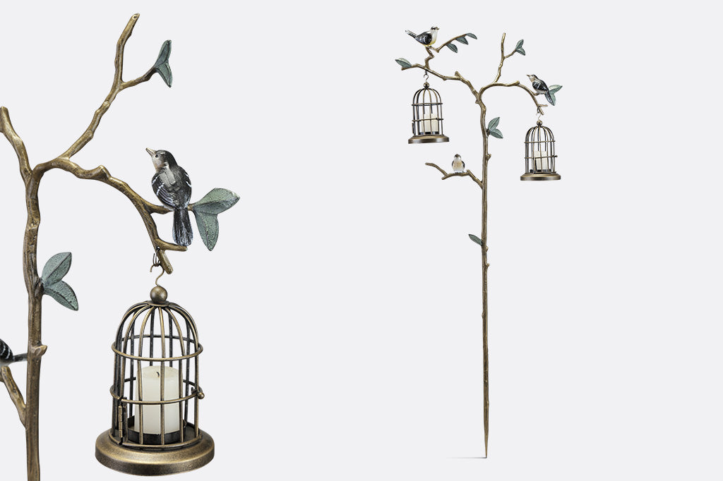 a staked garden accent with branch and leaf motif with songbirds. It supports two hanging candle lantern cages.