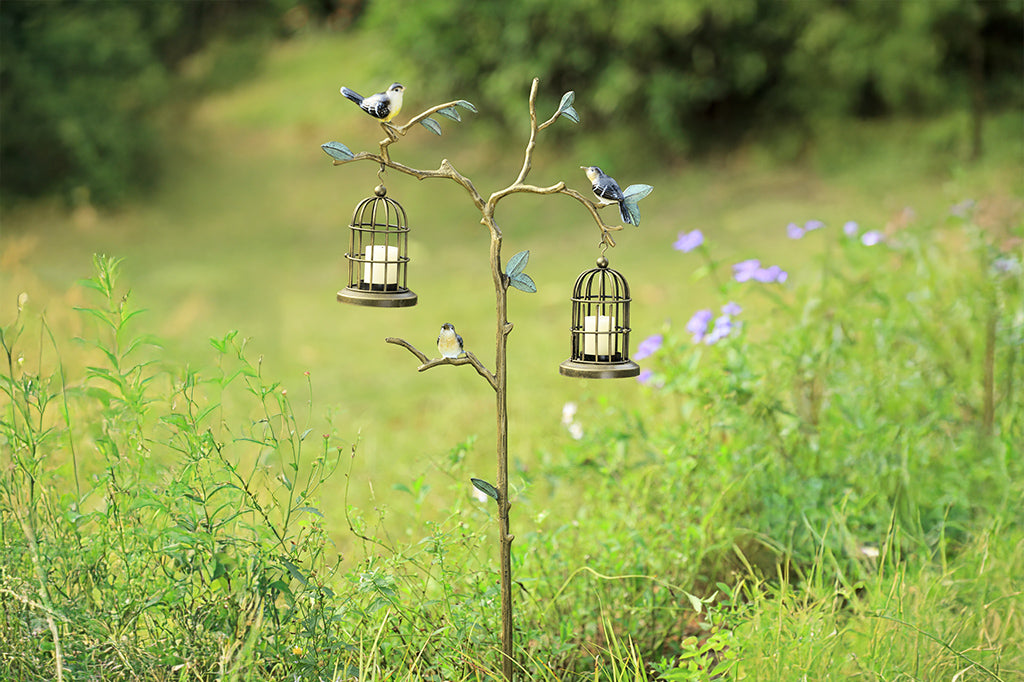 a staked garden accent shown amongst floral shrubbery. Branch and leaf motif with songbirds supports two hanging candle lantern cages.