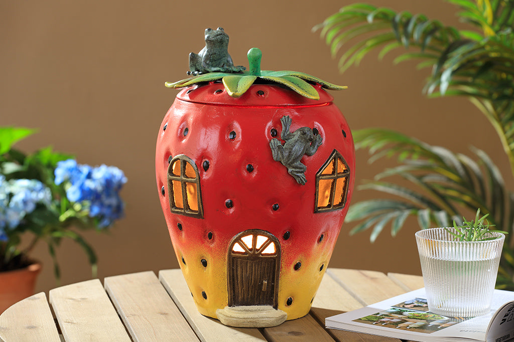 Strawberry House theme garden candle lantern with two frogs on top. Stem shaped lid comes off and candlelight shines through windows and door of strawberry house. Shown on oudoor patio table near book and beverage with potted plants in background.