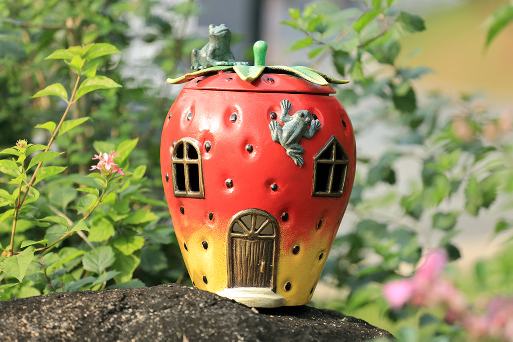 Strawberry House theme garden candle lantern with two frogs on top. Stem shaped lid comes off and candlelight shines through windows and door of strawberry house. Shown on rock in garden.