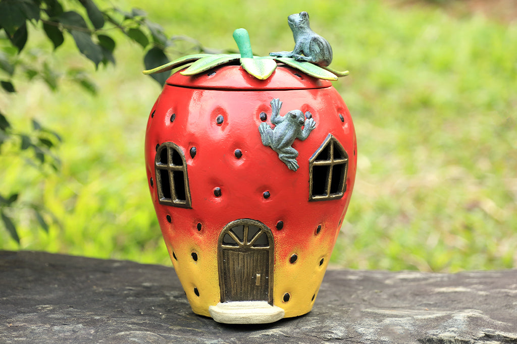 Strawberry House theme garden candle lantern with two frogs on top. Stem shaped lid comes off and candlelight shines through windows and door of strawberry house. Shown on rock in garden.