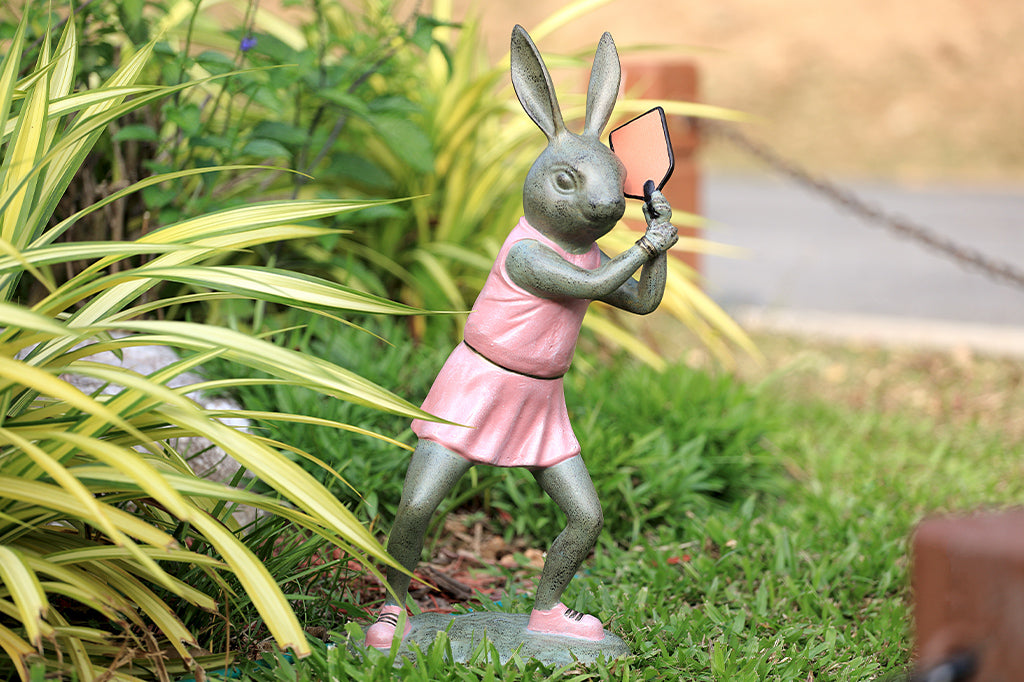 Rabbit garden sculpture with pickleball theme. She wears a pink dress, pink sneakers and holds a pink pickleball paddle in a garden setting