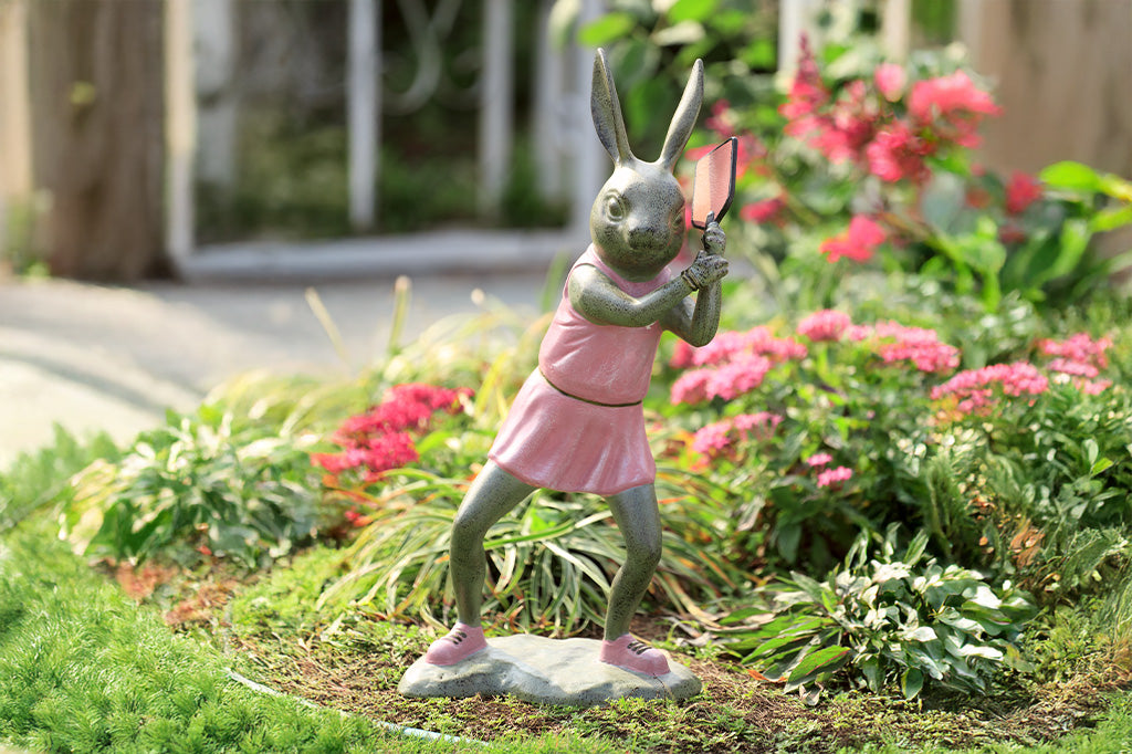 Rabbit garden sculpture with pickleball theme. She wears a pink dress, pink sneakers and holds a pink pickleball paddle in a garden setting