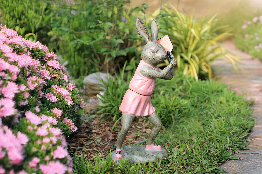 Rabbit garden sculpture with pickleball theme. She wears a pink dress, pink sneakers and holds a pink pickleball paddle in a garden setting