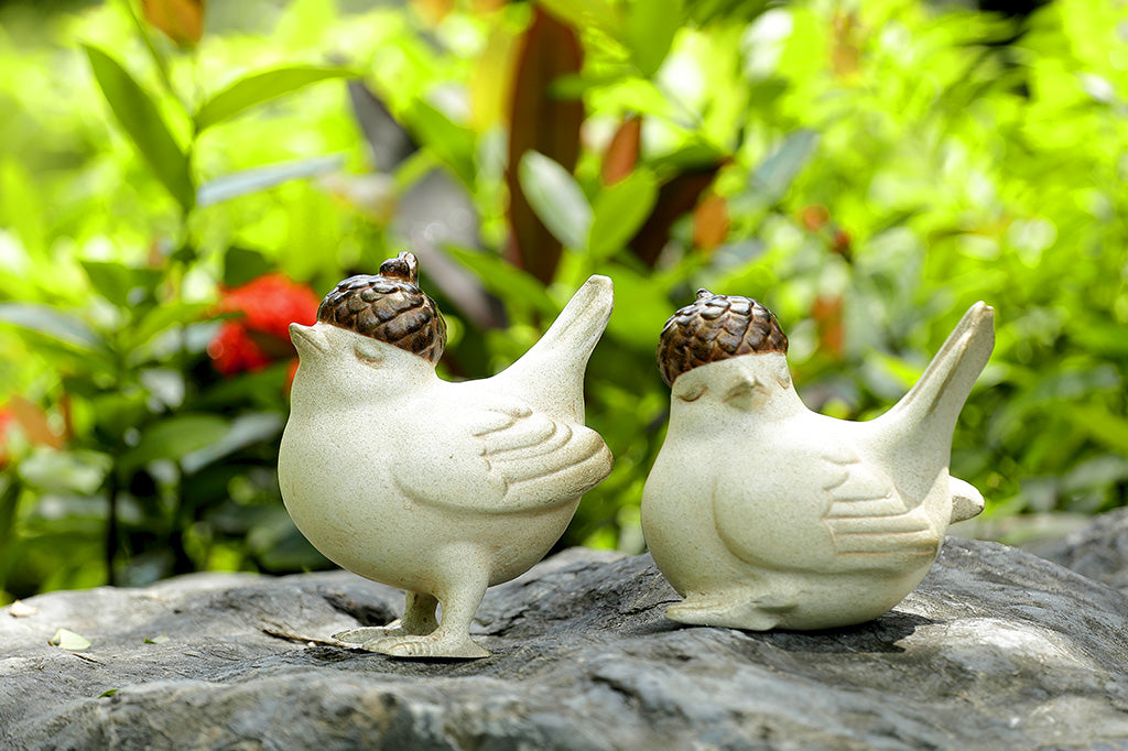 A pair of two birds, one standing, the other sitting, both wearing little acorn hats placed in a garden.
