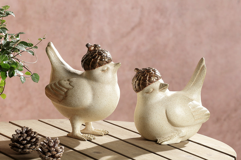 A pair of two birds, one standing, the other sitting, both wearing little acorn hats placed on a table with pinecone decor.