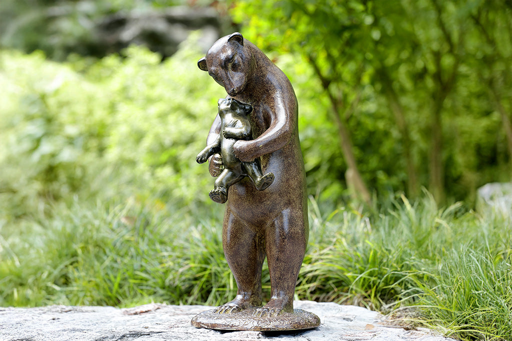 A brown sculpture of a bear holding a cub to hug them placed in a garden.