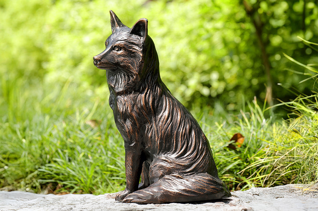 The side profile of a brown sculpture of a fox sitting, placed in a garden.