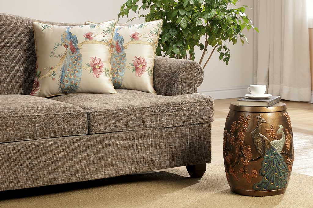 embroidered peacock on branch with peony flowers on champagne satin pillow shown on couch  in living room with peacock garden stool