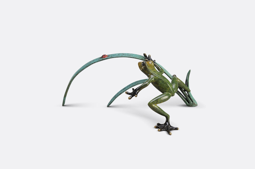 A green frog figurine on top of long blue-green leaves ready to pounce a lady bug.