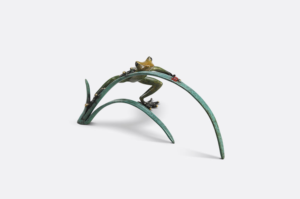 A green frog figurine on top of long blue-green leaves ready to pounce a lady bug.