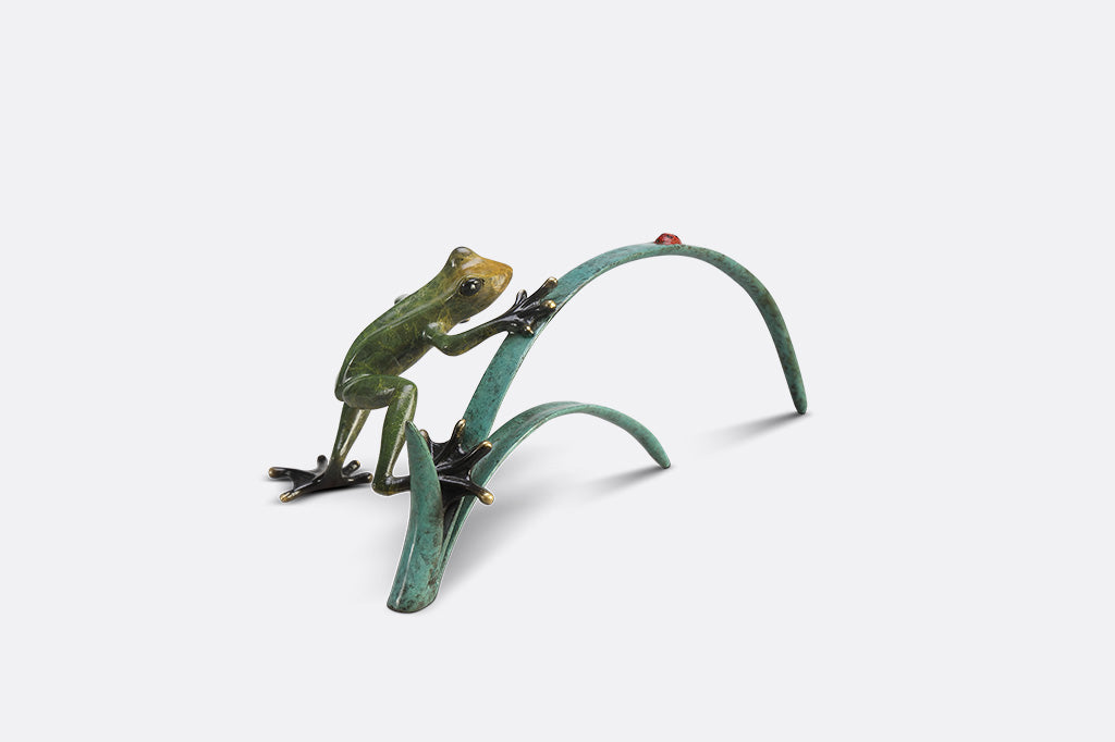 A green frog figurine on top of long blue-green leaves ready to pounce a lady bug.
