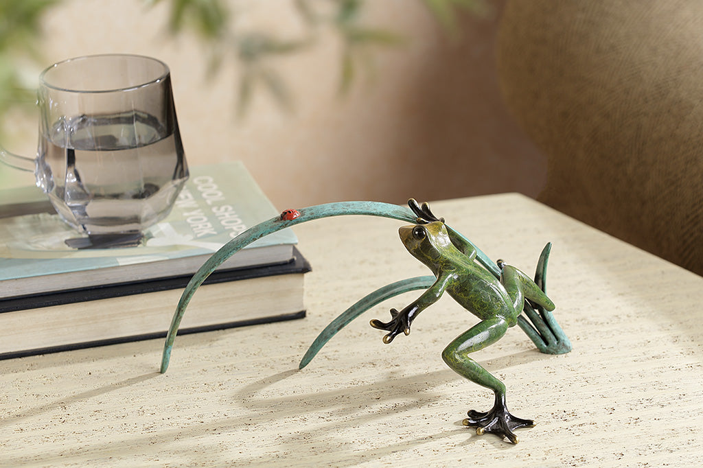 A green frog figurine on top of long blue-green leaves ready to pounce a lady bug.