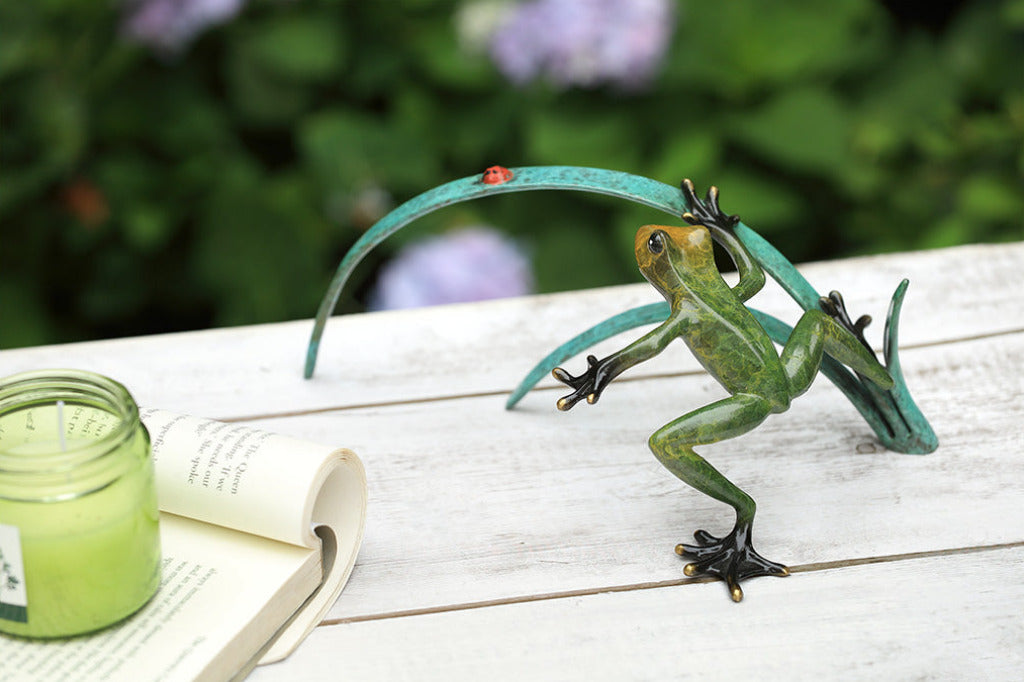 A green frog figurine on top of long blue-green leaves ready to pounce a lady bug.