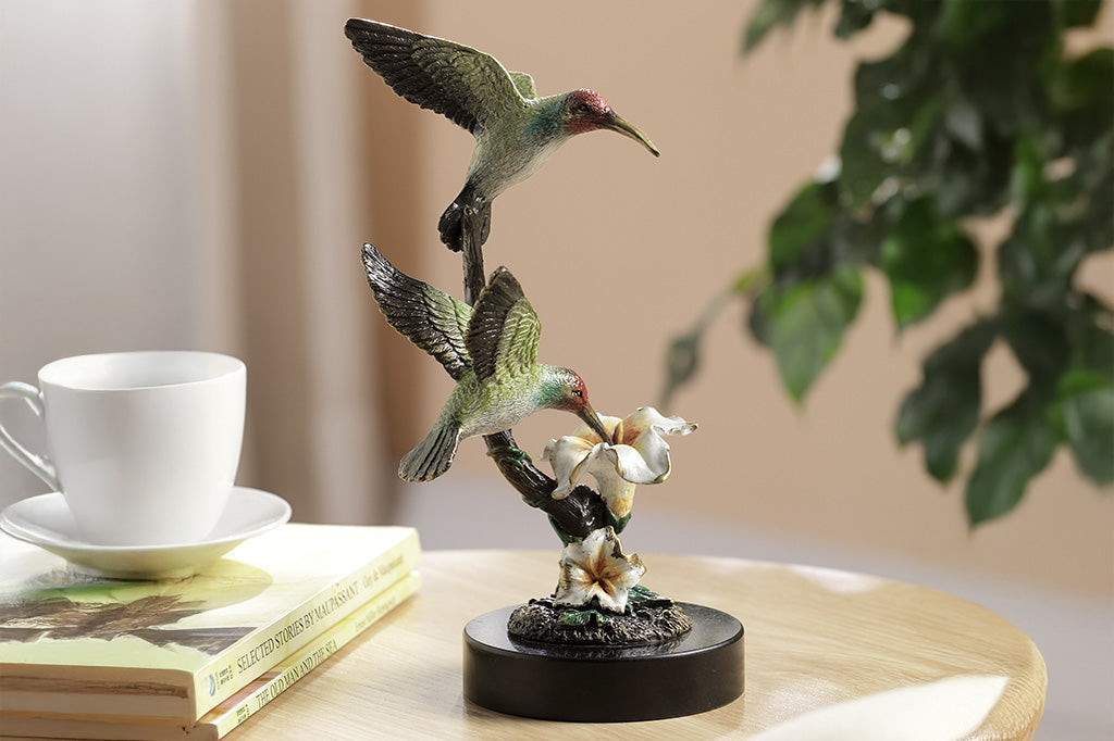 The side profile of a sculpture of two birds, one perched on a branch, and another taking a sip of nectar from a flower placed on a table.