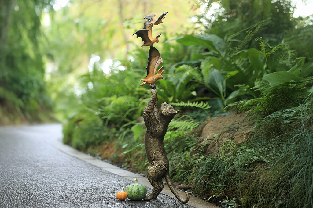 cast metal cat sculpture on back paw reaching/ playing with orange and black bats with two pumpkins by base of kitty hind paws set in garden scene 