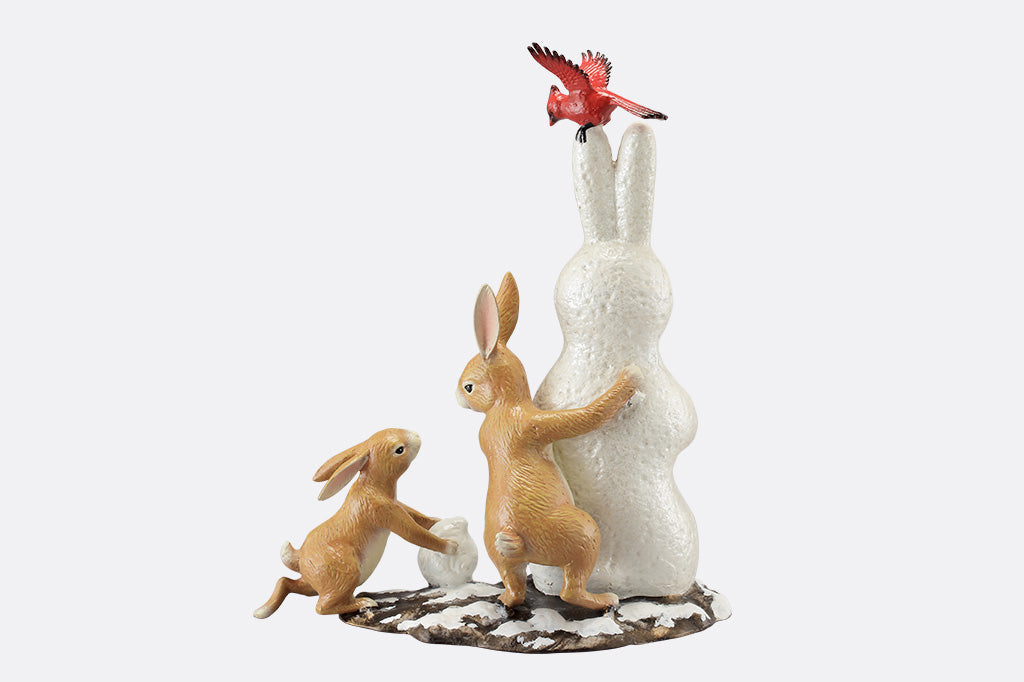 The back of a sculpture of a pair of two bunnies building a bunny shaped snow man, while a cardinal bird hovers above.