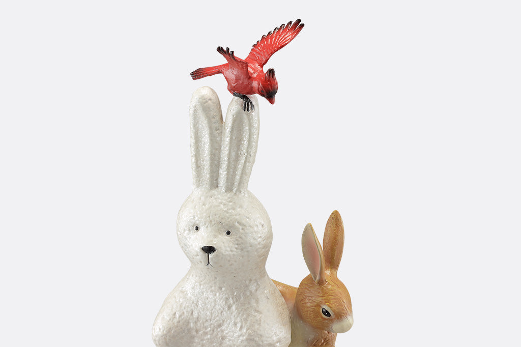 A close up of a sculpture of a pair of two bunnies building a bunny shaped snow man, while a cardinal bird hovers above.